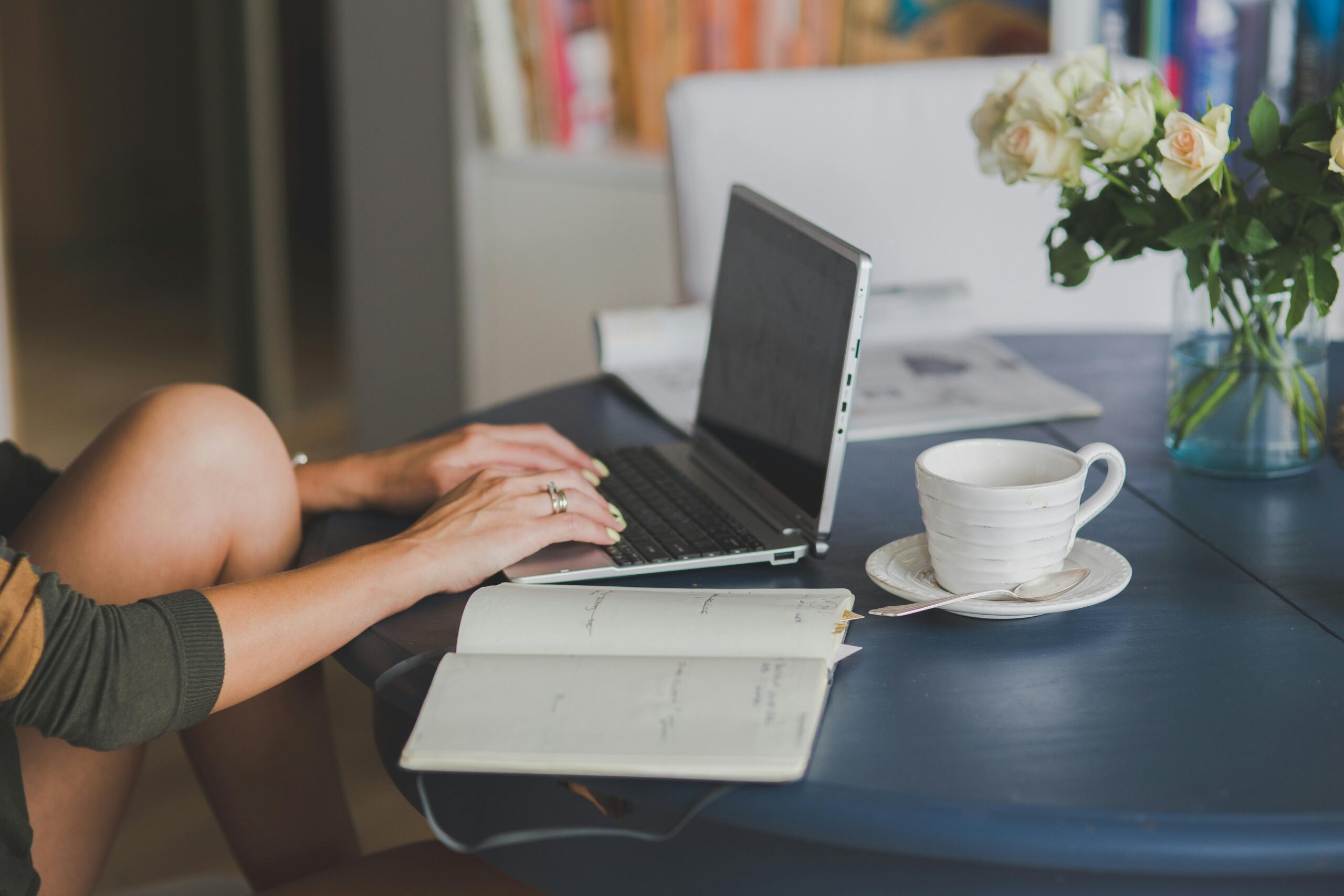 Woman working on a laptop from home with an open diary, coffee, and flower vase.